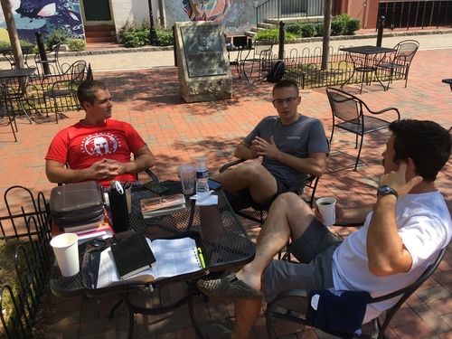 three men sitting at an outdoor table with Bibles and coffee